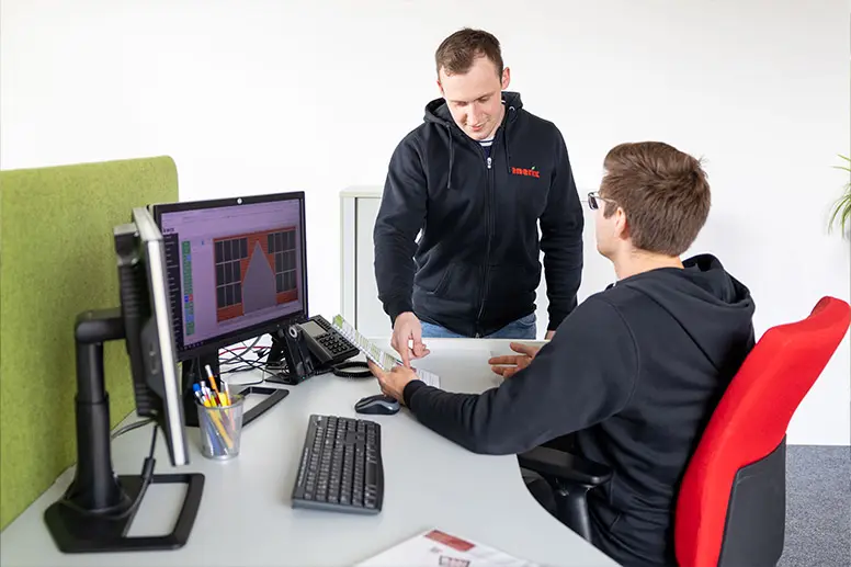 Ein Photovoltaik Projektleiter steht an einem PC und spricht mit einem Kollegen der auf einem Stuhl sitzt.