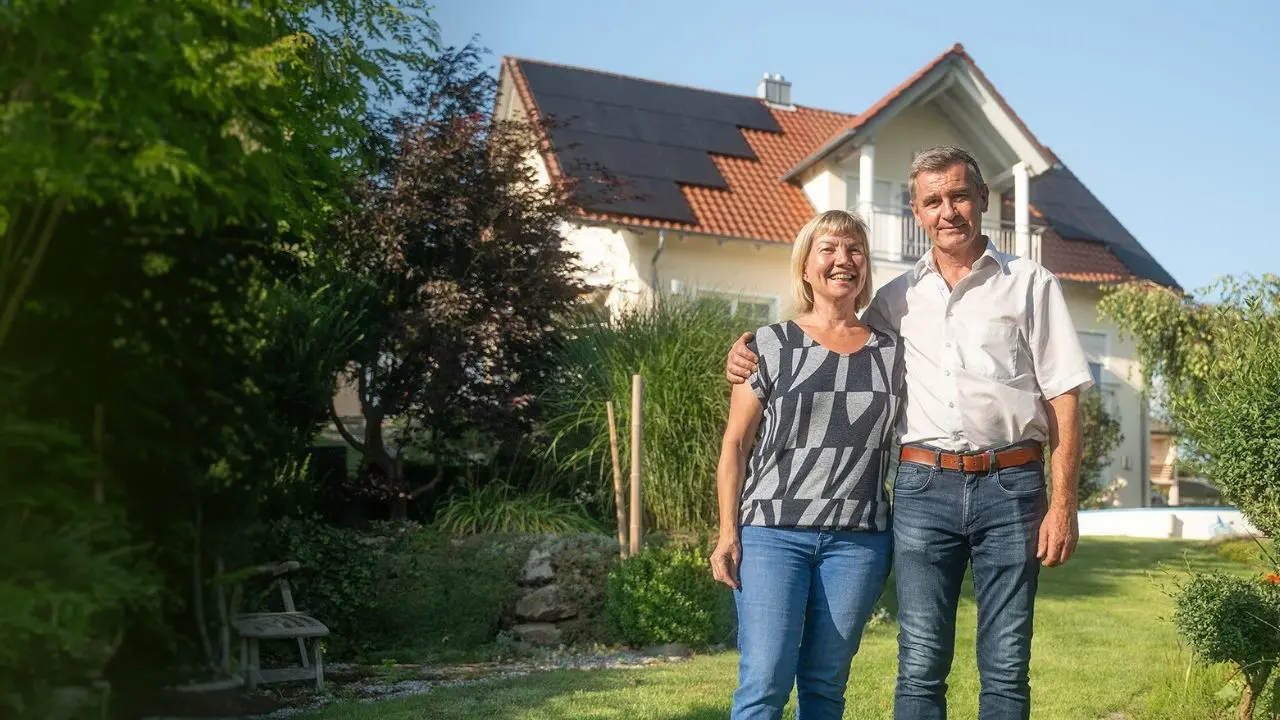 Ein glückliches Ehepaar mittleren Altern steht in ihrem Garten vor ihrem Haus mit einer neu montierten Photovoltaikanlage.