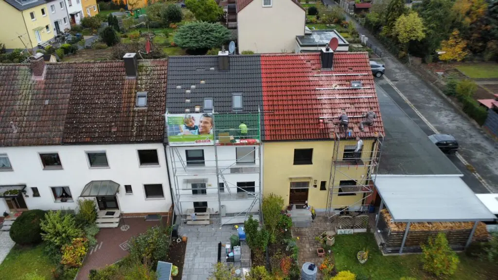 Zwei verschiedene Photovoltaikfirmen montieren gleichzeitig eine Photovoltaikanlage auf zwei nebeneinander stehenden Hausdächern. Eine Firma verwendet einen großen Fallschutzzaun, um ihre Monteure vor möglichen Abstürzen zu sichern. Die andere Firma hat hingegen nur ein zu kleines Gerüst.