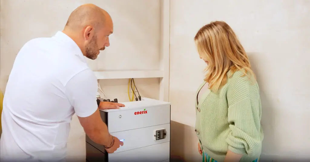 Ein Fachberater von enerix erklärt einer Frau einen Stromspeicher ohne PV-Anlage. Die beiden stehen im Keller vor dem angeschlossenen Gerät, während der Berater auf die Bedienoberfläche des Speichers zeigt.