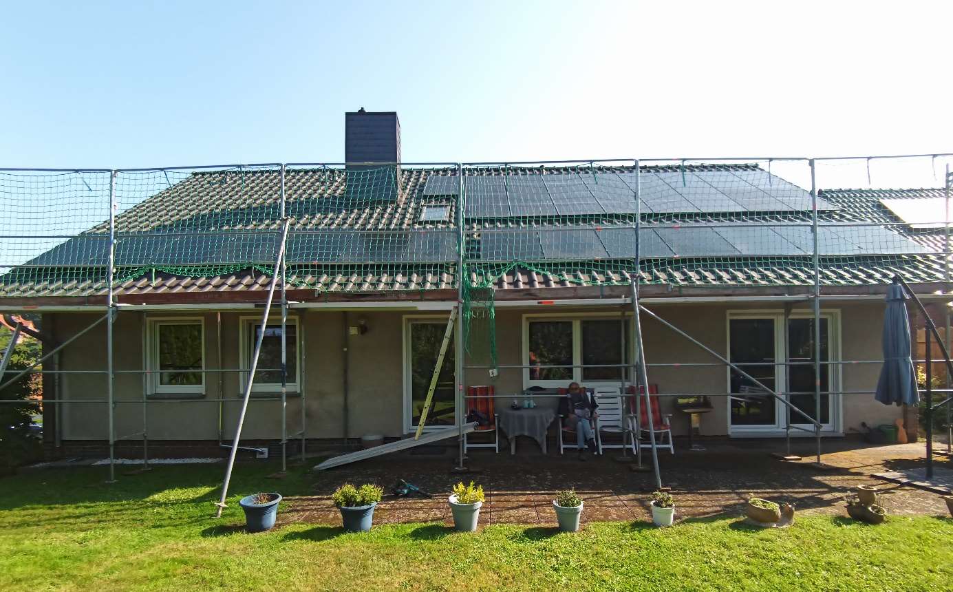 Ein Haus mit einer nicht fertiggestellten Photovoltaikanlage. Eine Person sitzt auf der Terrasse und das Gerüst steht immer noch um das Haus herum.