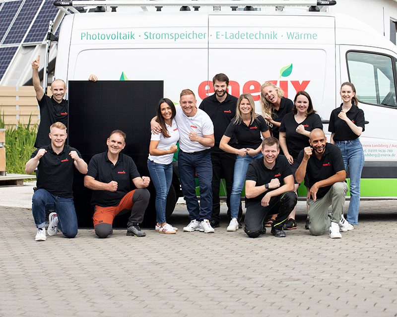 Das Team von enerix Landsberg Augsburg steht vor einem Montagefahrzeug von enerix und lacht motiviert. Im Hintergrund eine PV-Anlage auf einem Dach.