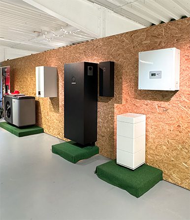 Eine Ausstellung mit verschiedenen Energiespeicher- und Photovoltaik-Komponenten an einer Wand mit Holzverkleidung. Zu sehen sind mehrere Batteriespeicher, ein Wechselrichter und eine Wärmepumpe, die auf kleinen Podesten mit grünem Teppich platziert sind.