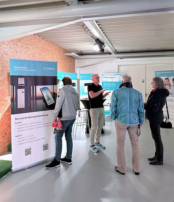 ine Beratungssituation in einer Ausstellung für Energiespeicherlösungen. Ein Berater in schwarzem Shirt spricht mit drei interessierten Besuchern, die sich über Photovoltaik und Batteriespeicher informieren. Im Vordergrund steht ein Werbebanner der Firma Fenecon mit Informationen zur „Home Serie“ für smarte Heimspeicherlösungen. Im Hintergrund sind weitere Informationsstände und Displays mit technischen Details zu sehen. 