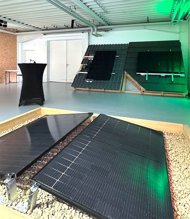 Eine Ausstellung mit verschiedenen Photovoltaik-Modellen. Im Vordergrund sind zwei Solarmodule auf einem Kiesbett montiert. Im Hintergrund befinden sich zwei geneigte Dachmodelle mit installierten Solarpaneelen, die unterschiedliche Montagesysteme zeigen. 