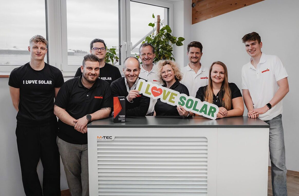 Das Team von enerix Süedschwarzwald steht gemeinsam hinter einem M-TEC Wärmepumpen-Innengerät in einem modernen Büro. Neun Personen – Frauen und Männer – tragen enerix- oder „I LOVE SOLAR“-Shirts und lächeln in die Kamera. Zwei Personen in der Mitte halten ein Schild mit der Aufschrift „I ❤️ LOVE SOLAR“ sowie eine Trophäe in der Hand. Im Hintergrund sind große Fenster und grüne Pflanzen zu sehen.