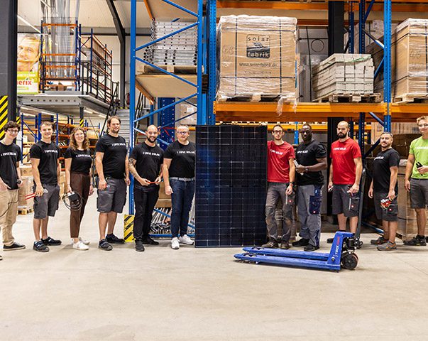Ein Team von Elektrikern, Solarteuren und Dachdeckern steht in einer Lagerhalle. Zwei der Personen halten ein aufgestelltes Solarmodule in den Händen