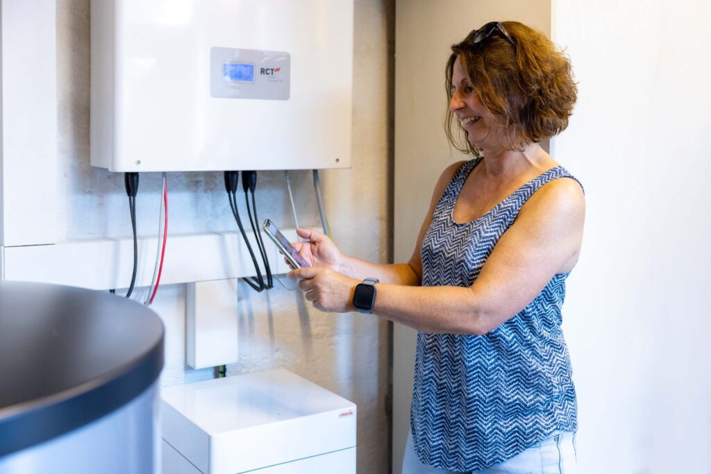 Eine Frau steht mit einem Handy vor dem nachgerüsteten PV Speicher und freut sich über die Einsparung. 