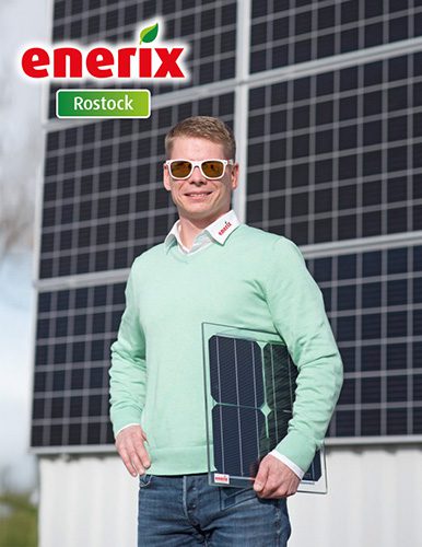 Der Geschäftsinhaber Björn Schumacher mit Sonnenbrille und grünem Pullover steht vor einer großen Photovoltaikanlage und hält ein kleines Solarmodul in der Hand. Er trägt ein Hemd mit dem Enerix-Logo am Kragen und lächelt in die Kamera. Oben links im Bild ist das enerix Rostock-Logo zu sehen.