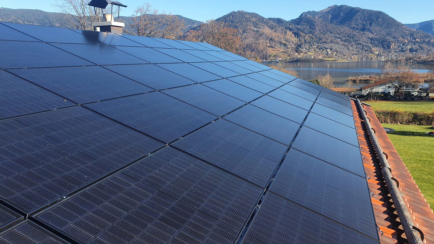 Eine große Photovoltaikanlage auf einem roten Ziegeldach mit einem großen See im Hintergrund bei gutem Wetter.