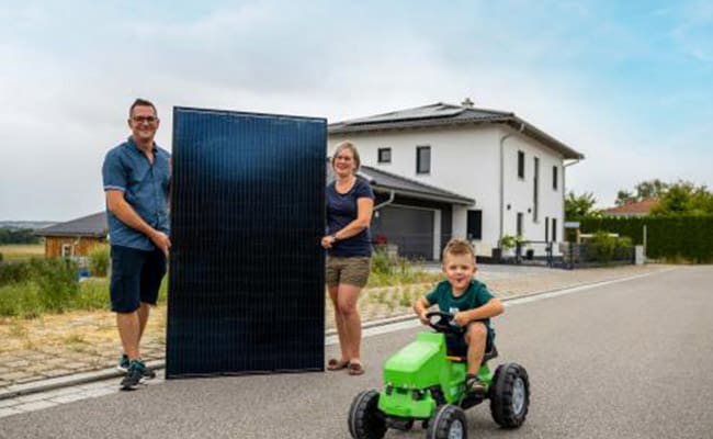 Ein Ehepaar hält ein Photovoltaikmodul und steht vor ihrem Haus mit einer neu montierten Photovoltaikanlage auf dem Dach. Der Sohn fährt auf einem Spielzeug Bulldog in das Bild und trägt ein enerix Solarstromhelden T-Shirt.