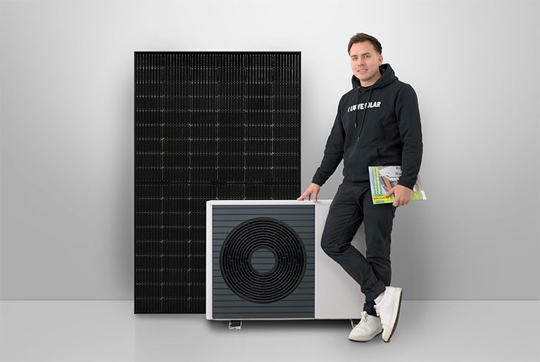 Eine Person steht vor einer Wärmepumpe und einem Photovoltaikmodul in der Hand.