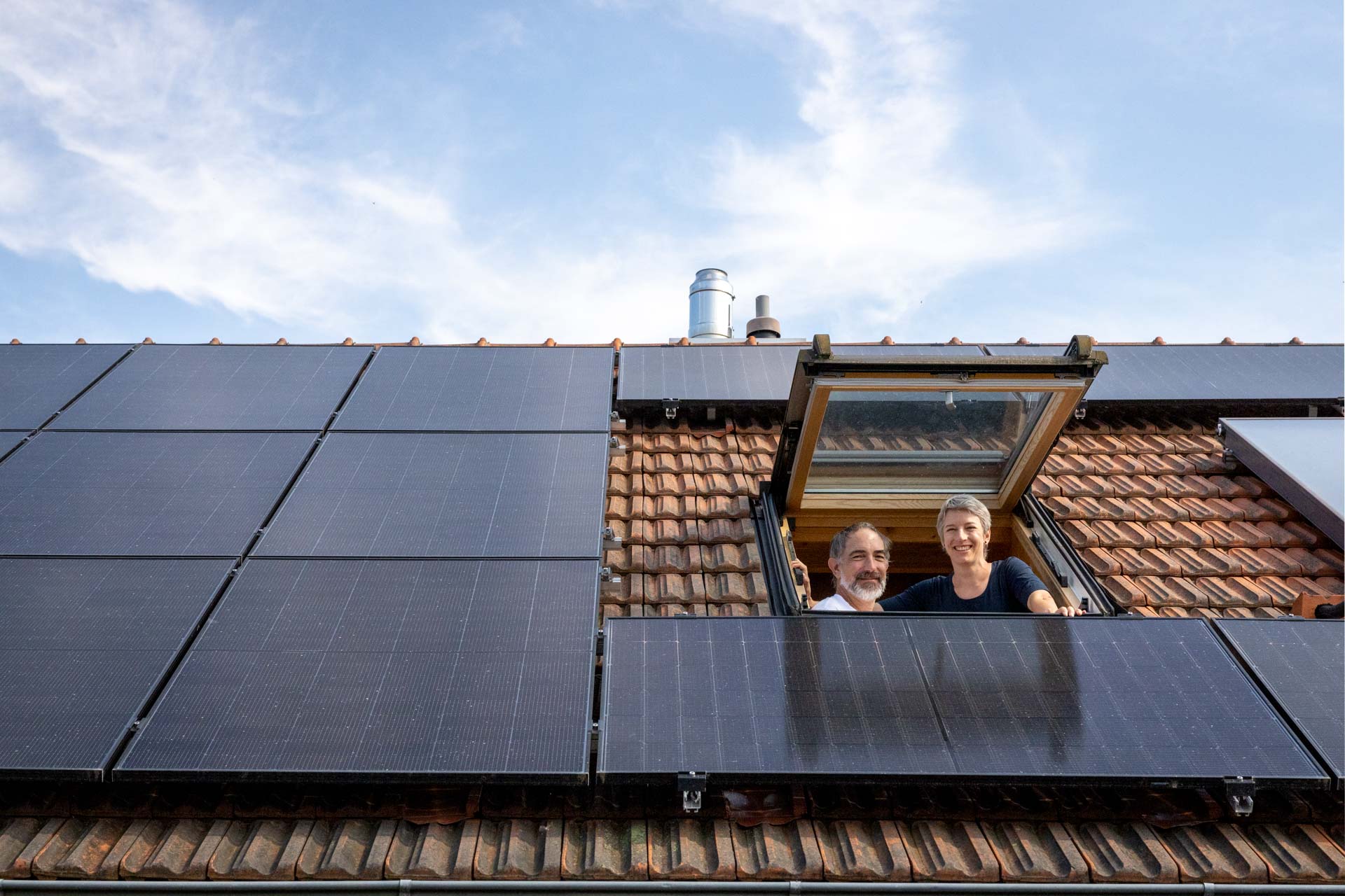 Ein rotes Hausdach mit einer Photovoltaikanlage und einem Dachfenster aus dem ein Ehepaar herausschaut.
