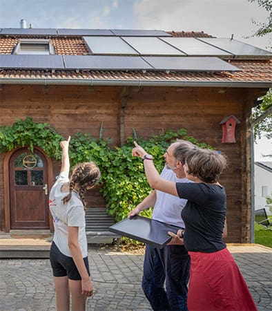 Eine Familie steht vor ihrem Haus, hält ein kleines Photovoltaikmodul in der Hand und zeigt auf die große Photovoltaikanlage auf ihrem Hausdach.