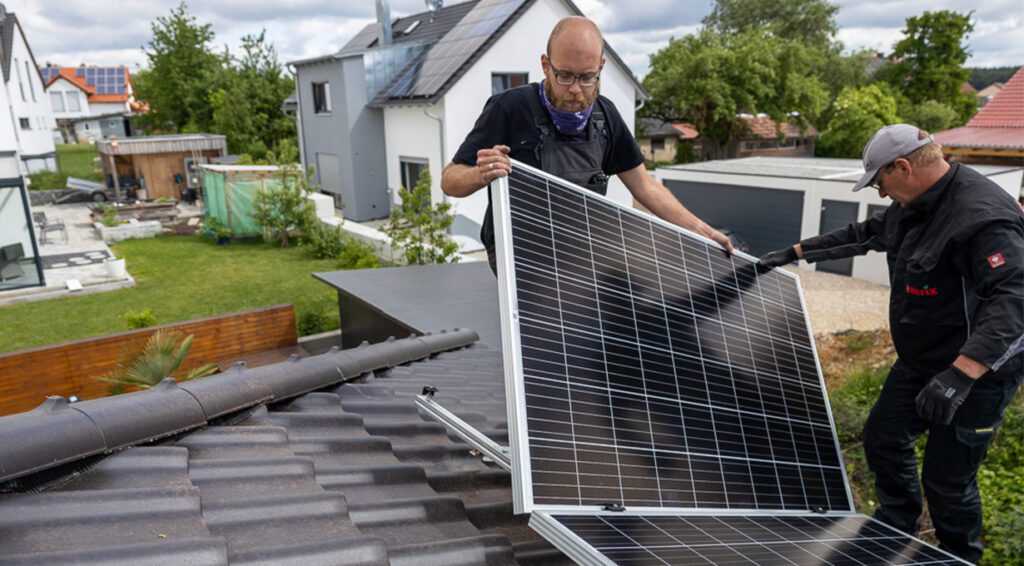 Zwei Monteure von enerix Stuttgart montieren ein Photovoltaikmodul auf einer Unterkonstruktion auf einem grauen Hausdach.