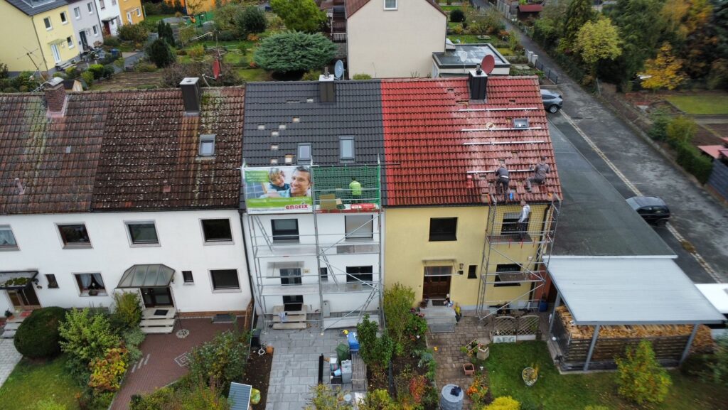 Zwei verschiedene Photovoltaikfirmen montieren gleichzeitig eine Photovoltaikanlage auf zwei nebeneinander stehenden Hausdächern. Eine Firma verwendet einen großen Fallschutzzaun, um ihre Monteure vor möglichen Abstürzen zu sichern. Die andere Firma hat hingegen nur ein zu kleines Gerüst.