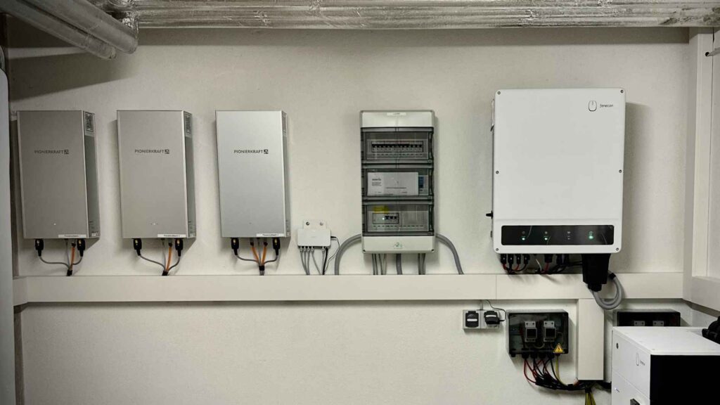 Drei Pionierkraft Mieterstromkästen sind in einem Keller neben einem Stromkasten und einem Wechselrichter montiert.