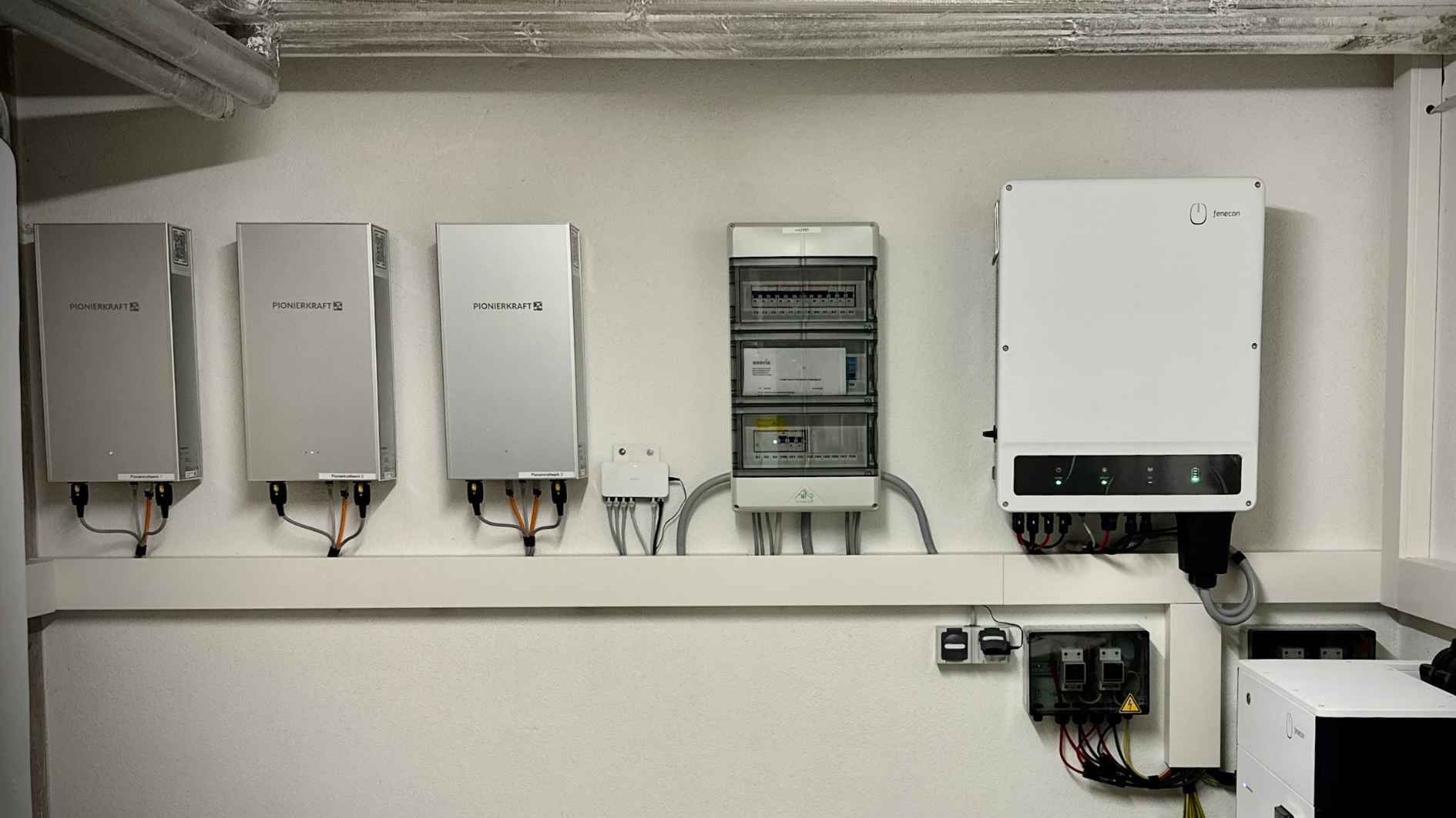 Drei Pionierkraft Mieterstromkästen sind in einem Keller neben einem Stromkasten und einem Wechselrichter montiert.