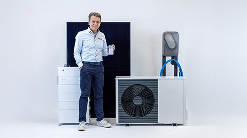 Der Geschäftsführer von enerix Peter Knuth steht vor einem Speicher, einem Photovoltaikmodul, einer Wärmepumpe und einer Wallbox und präsentiert auf einem Smartphone den Energiemanager enerixControl.