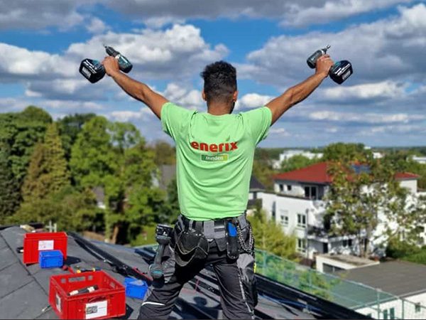 Ein Solarmonteur steht auf einem Hausdach und hält zwei Schraubendreher in die Luft.