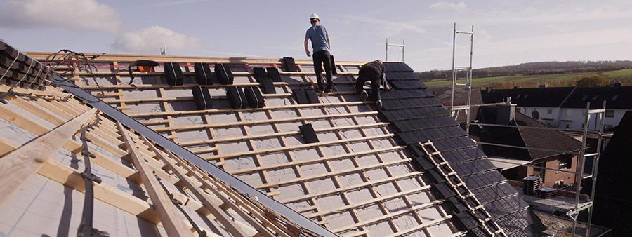Zwei Handwerker installieren Solardachziegel auf einem geneigten Dach, das teilweise bereits eingedeckt ist; professionelle Montage als Teil einer Dachsanierung.
