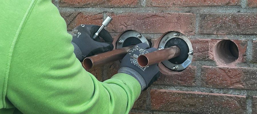 SHK-Meister bei der Installation einer Wärmepumpe: Kupferleitungen ragen durch eine gebohrte Hauswand – Montage durch enerix Weser-Ems.
