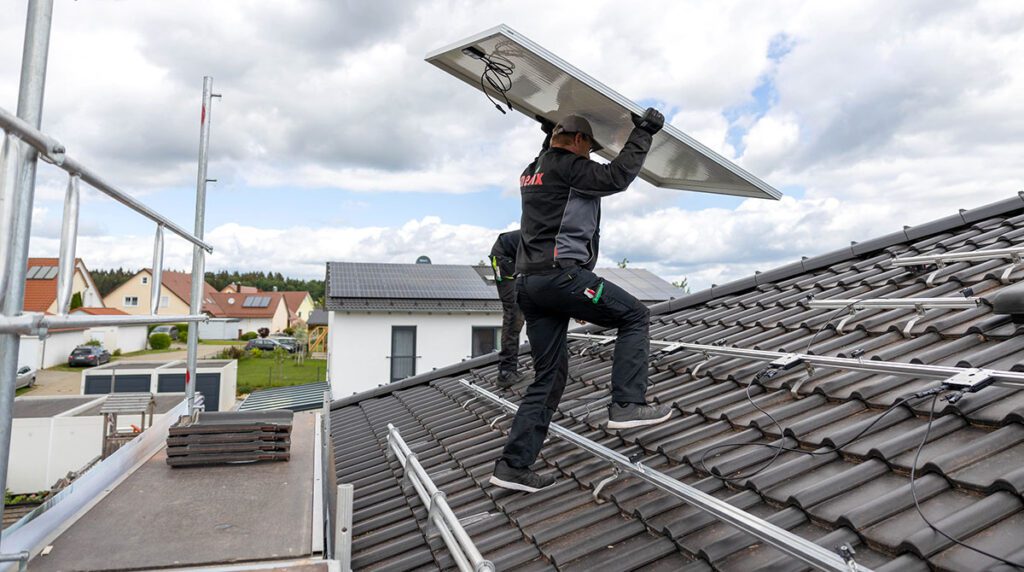 Handwerker verschraubt ein Photovoltaikmodul auf dem Dach, Montage mit Unterkonstruktion sichtbar.