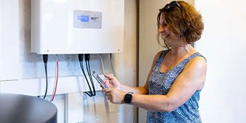 Eine Frau steht vor einem RCT Wechselrichter und schaut auf ihr Smartphone