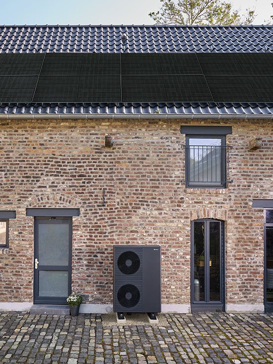 Eine schwarze doppelte Wärmepumpe von Vaillant steht vor einem renovierten Ziegelhaus. Auf dem Dach sind Solarmodule zu sehen.