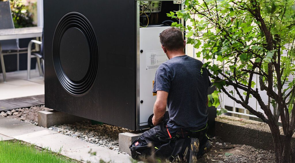 Ein Mann kniet vor einer Wärmepumpe und hat Werkzeug in der Hand