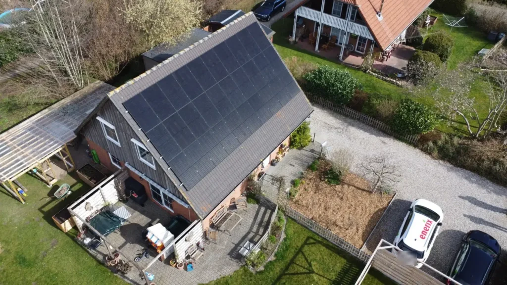 Luftaufnahme eines Backsteinhauses mit schwarzer Photovoltaikanlage auf dem Satteldach, Garten und E-Auto in der Einfahrt